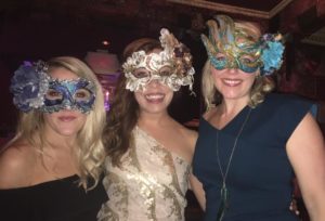 Three smiling women in fancy masquerade masks look at the camera