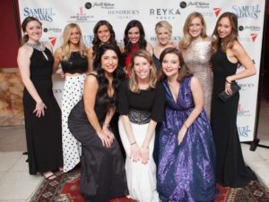 A group of women in formal gowns pose smiling in front of a banner with event sponsor logos at a past Junior League of Boston Charity Gala