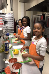 Three adolescent girls stand along a counter wearing orange aprons and chopping vegetables. They are participants in one of the Junior League of Boston community programs supported by your donation