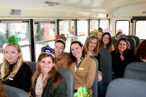 Smiling women wearing party hats are seated on a bus. The image shows Junior League members touring community programs throughout the city of Boston
