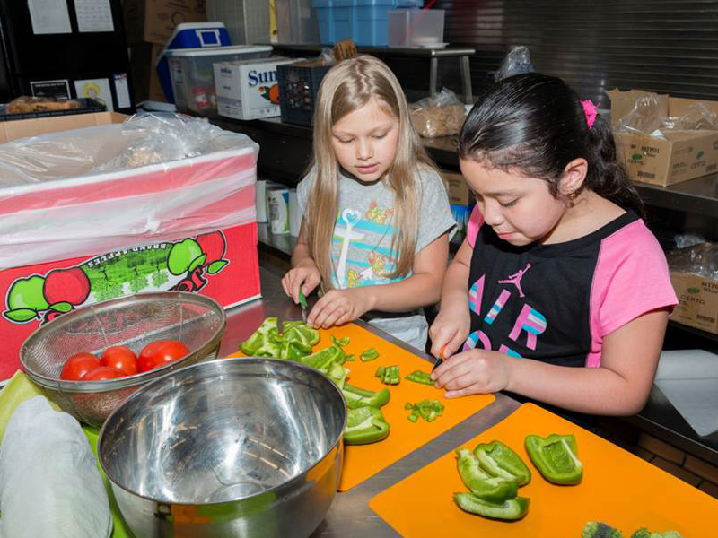 Two young girls cut green bell peppers on cutting boards as part of a Kids in the Kitchen session