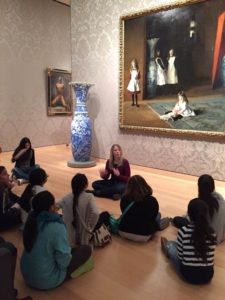 A woman sits cross-legged on the ground in an art museum in front of a large painting, gesturing with her hands. A group of adolescent girls sit on the floor around her and watch